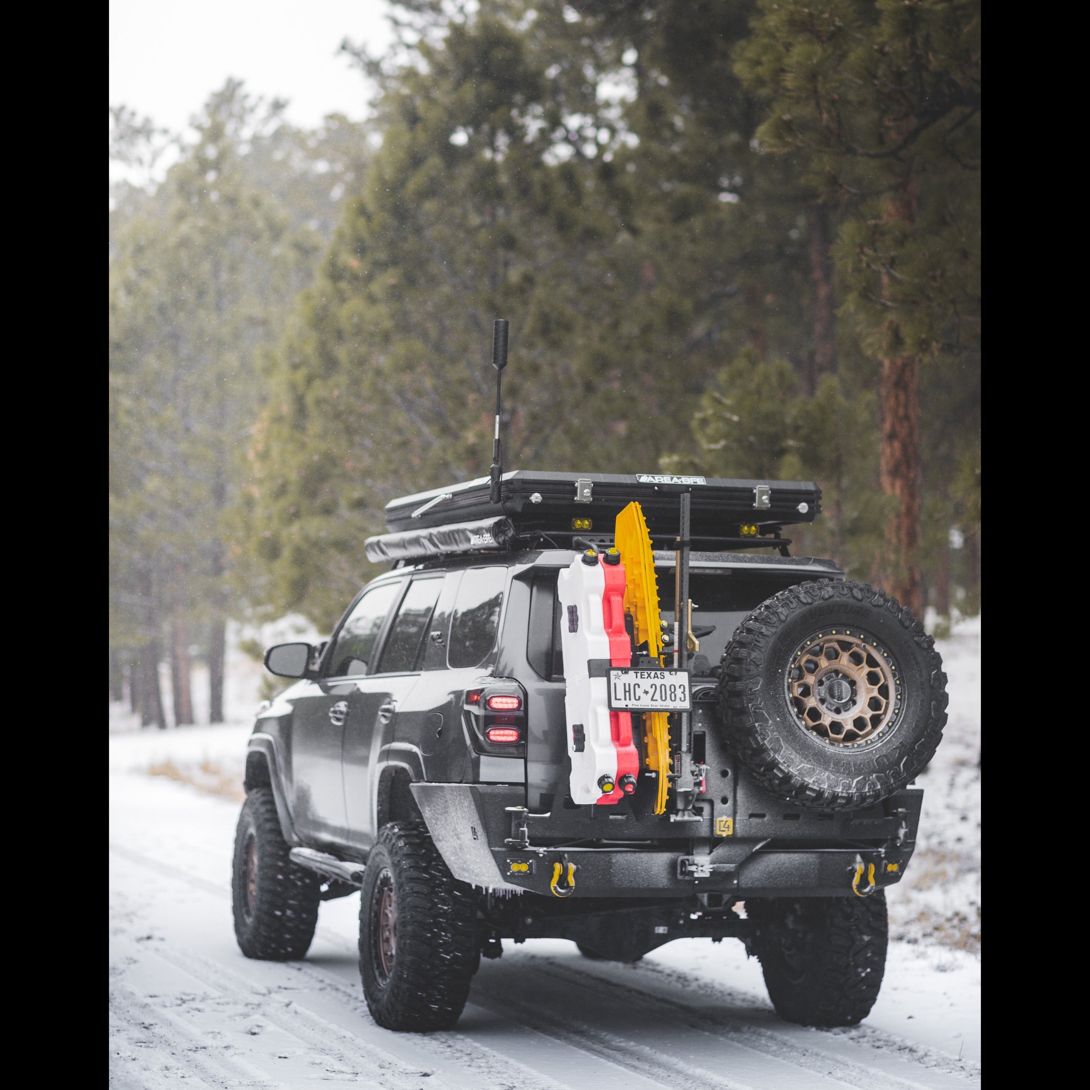 Grey 4Runner driving a mountain trail in the snow, equipped with a rooftop tent, awning, GMRS radio antenna and the C4 Fabrications steel rear bumper with dual swing-outs. 
