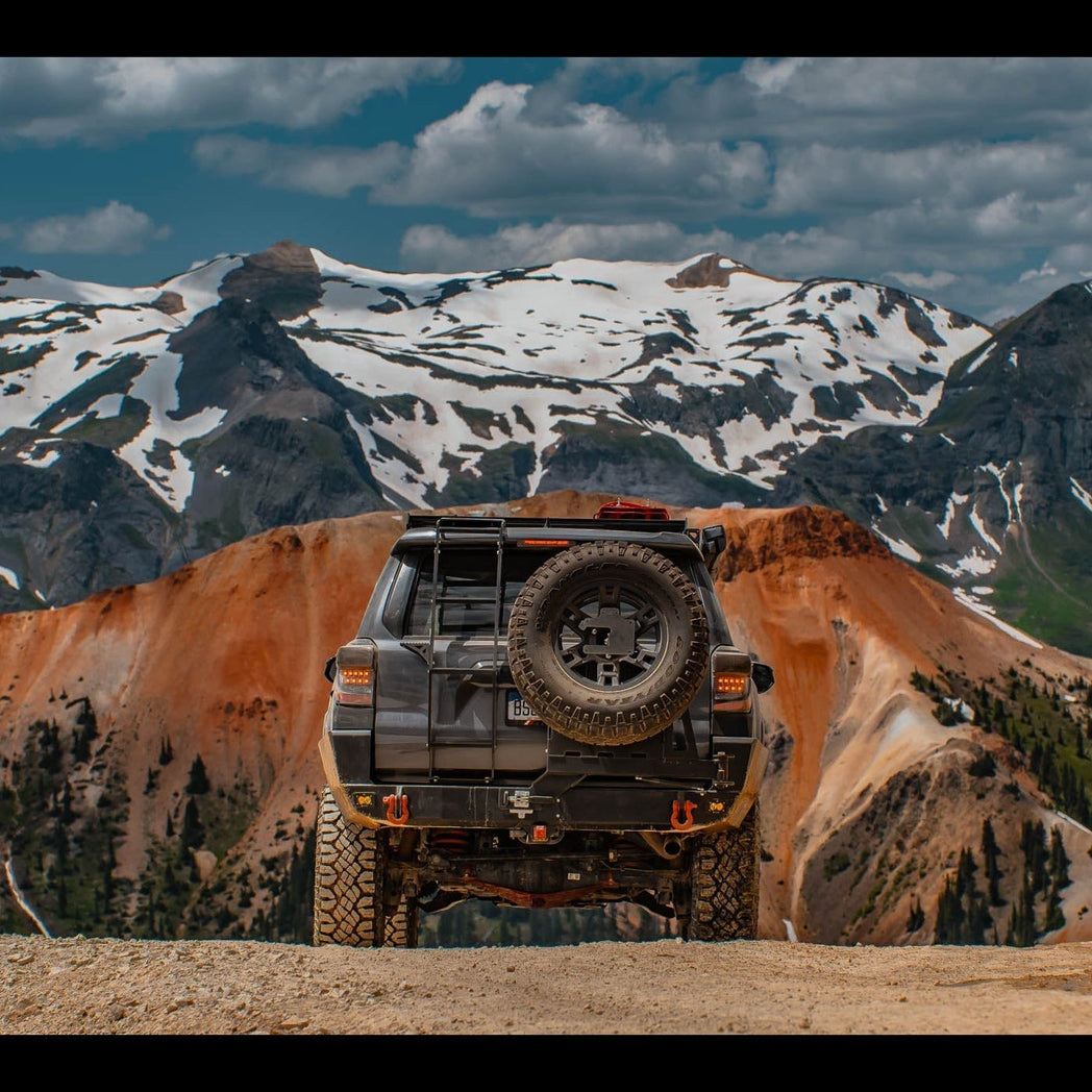 Grey 4Runner on a trail with snowcapped peaks in the background, equipped with a C4 single half swing Overland bumper with spare tire carrier and the Summit hatch ladder for accessing the gear secured to its roof.