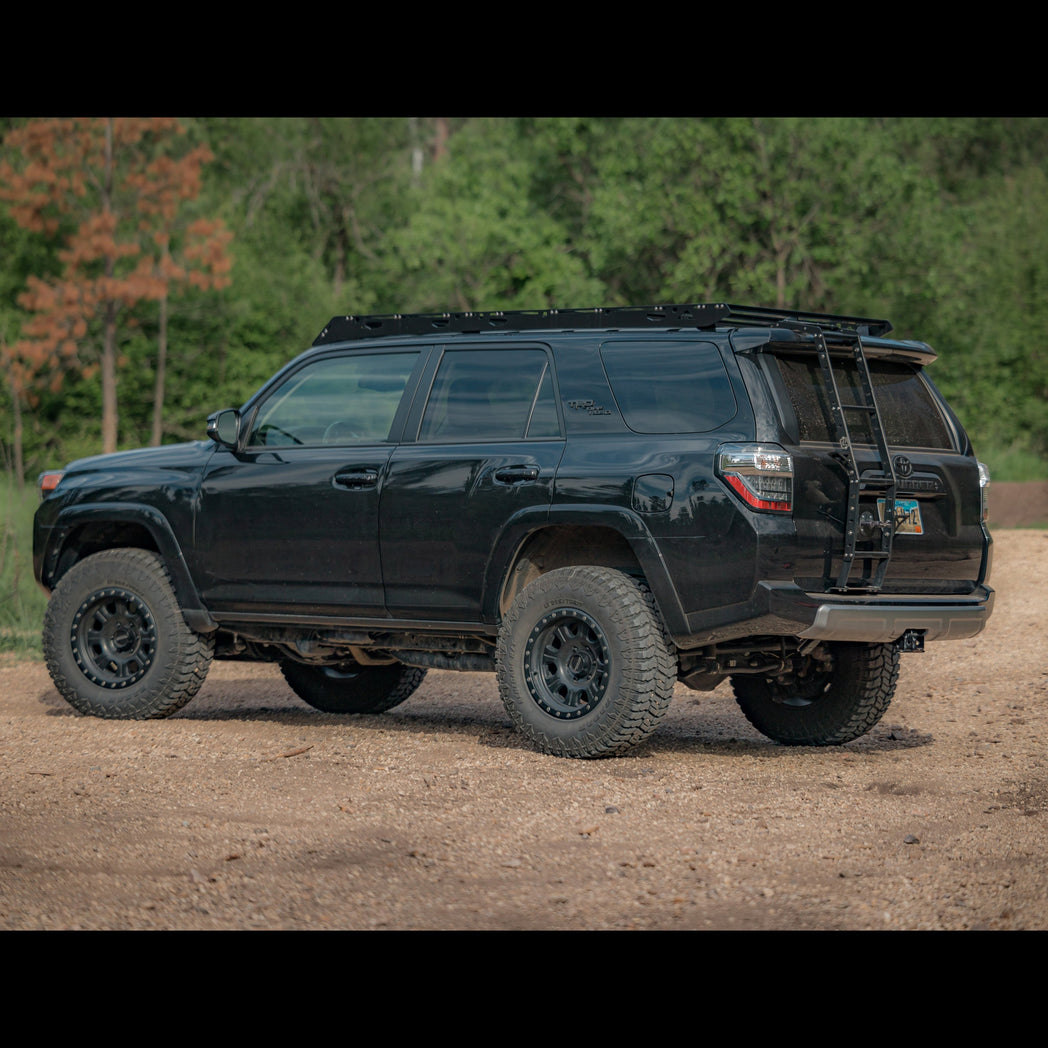 Wide view from the driver's rear showing a black 4Runner on 33" tires on a dirt road with trees in the background.