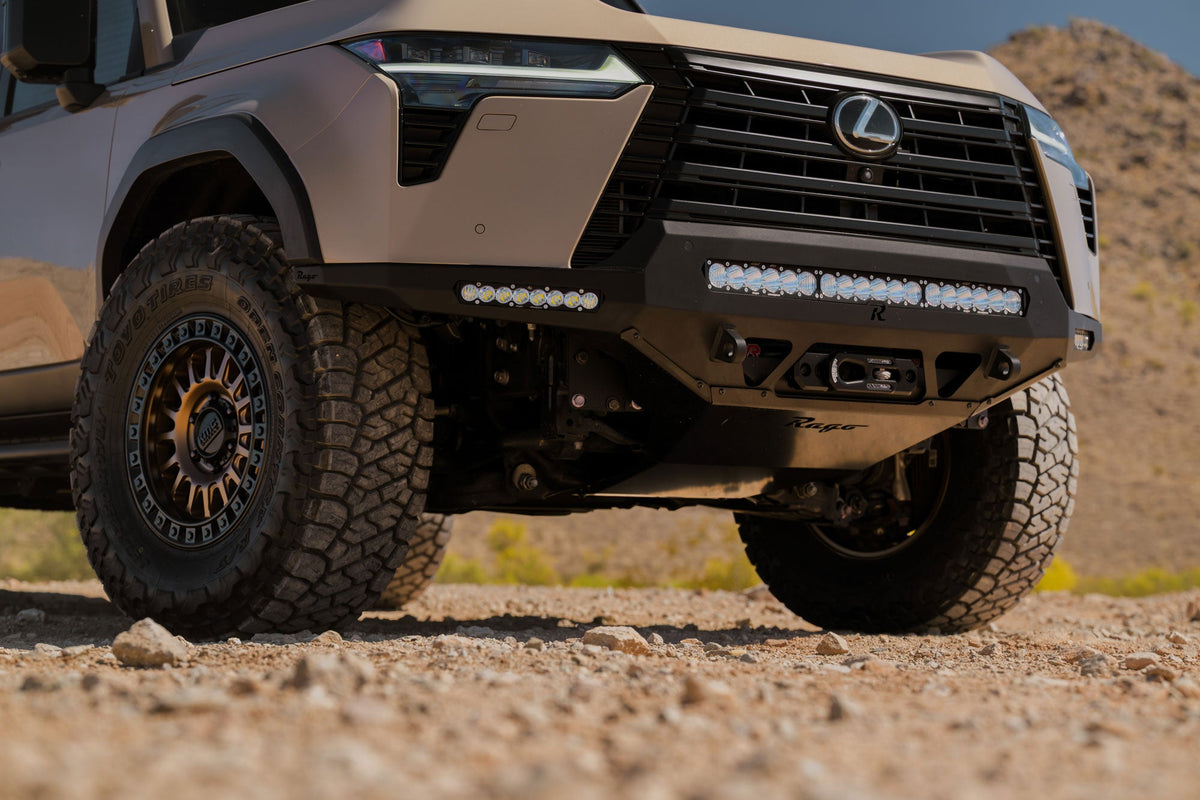 Front view of a Lexus off-road vehicle on a rocky terrain. It's equipped with Rago Fabrication's front bumper and skid plate, with added winch and light bars.