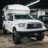 C4 Overland plate bumper on a lifted and modified white Toyota Tundra with a fiberglass slide-in camper installed. 