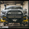 White Tacoma on a lift with the wheels off and zoomed in on the C4 Hybrid winch bumper with full compliment of lighting.